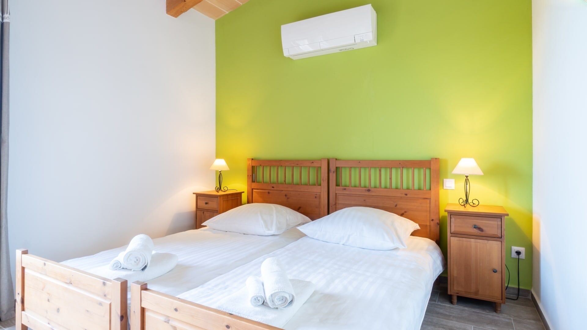 Un lit bien fait, avec du linge de lit blanc et des serviettes pliées, une tête de lit en bois, des tables de chevet avec des lampes et un appareil de climatisation fixé au mur.