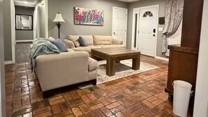 A living room with a sectional sofa, a coffee table, and a patterned rug. There are two doors, a lamp, and a painting on the wall.