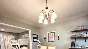 A dining area with a wooden table and chairs, a chandelier, a bunk bed, and a kitchen area with shelves.