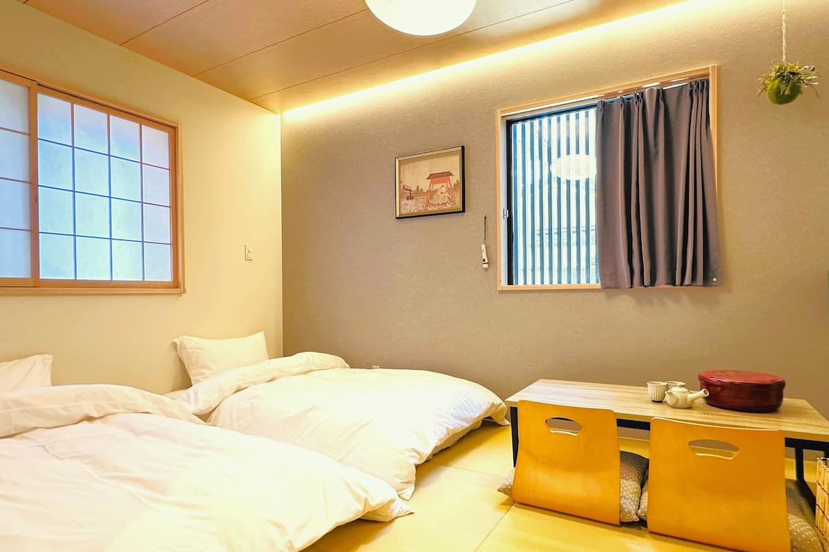 Une chambre avec deux lits, une petite table sur laquelle se trouve un pot rouge, et une fenêtre avec des rideaux.