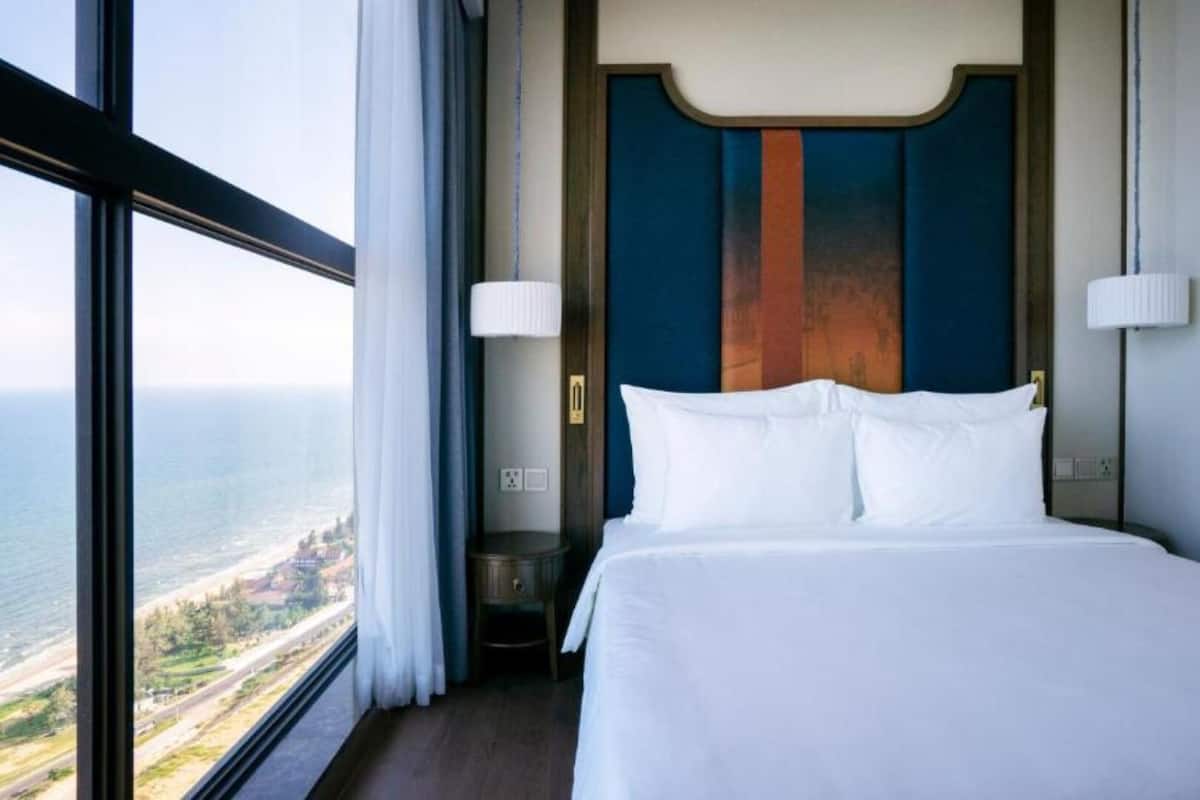 Camera d'albergo con una grande finestra che si affaccia sull'oceano e su una strada. La stanza presenta un letto con lenzuola bianche, una testiera in legno e due lampade ai lati del letto.