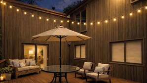 Un patio al aire libre con una mesa redonda de vidrio, muebles de mimbre e iluminación con luces colgantes.