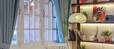 A room with two white rocking chairs, a wooden floor, a window with curtains, and a shelf with decorative items.