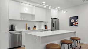 Una cocina moderna con armarios blancos, lavavajillas de acero inoxidable, un refrigerador y una isla central con taburetes.