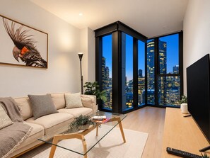 A modern living room with a sofa, coffee table, and a large window offering a cityscape view. - Skyline Suites | Stunning City Views, Gym & Pool (Southbank)
