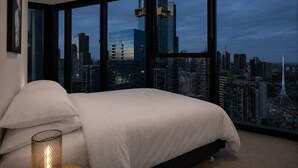 A bedroom with a large bed, a bedside lamp, and a view of a cityscape through large windows. - Skyline Suites | Stunning City Views, Gym & Pool (Southbank)