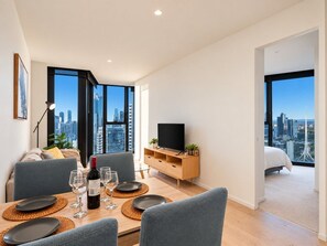 A modern living room with a dining area, a flat-screen TV, and a city view. - Skyline Suites | Stunning City Views, Gym & Pool (Southbank)