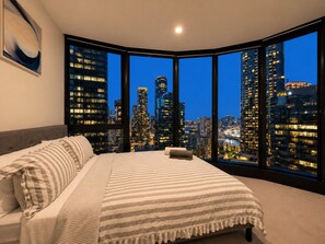 A bedroom with a large bed, a view of a cityscape at dusk, and a painting on the wall. - Skyline Suites | Stylish Corner Suite, City Views (Southbank)