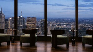 Een kamer met grote ramen en uitzicht op de stad, een ronde tafel en twee groene fauteuils.