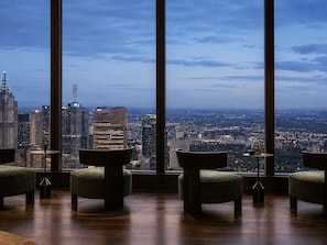 A room with large windows offering a cityscape view, a round table, and two green upholstered chairs. - Skyline Suites | Cosy Study Suite, Australia 108 (Southbank)
