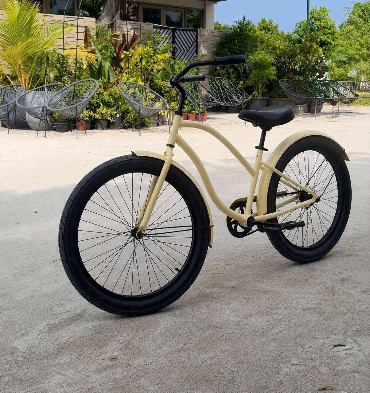 Una bicicleta estática con asiento y manubrio negros, sobre una superficie de concreto, con fondo de follaje y mobiliario exterior.