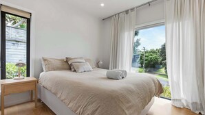 A bedroom with a bed, a nightstand, and a window with curtains. - Up Above The Crescent (Waihi Beach)