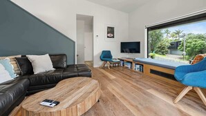 A modern living room with a black leather sofa, a wooden coffee table, a blue armchair, and a TV on a wooden stand. - Up Above The Crescent (Waihi Beach)