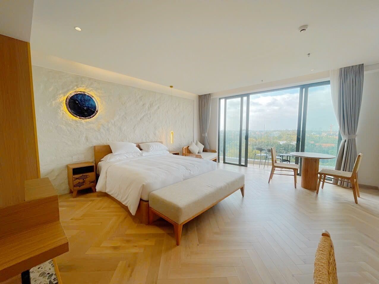 Une chambre moderne avec un grand lit, un banc en bois, un miroir rond et une vue sur la ville grâce à de grandes fenêtres.