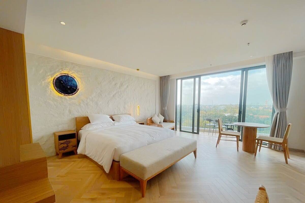 Un dormitorio moderno con una cama grande, un banco de madera, un espejo redondo y vistas de la ciudad a través de amplios ventanales.