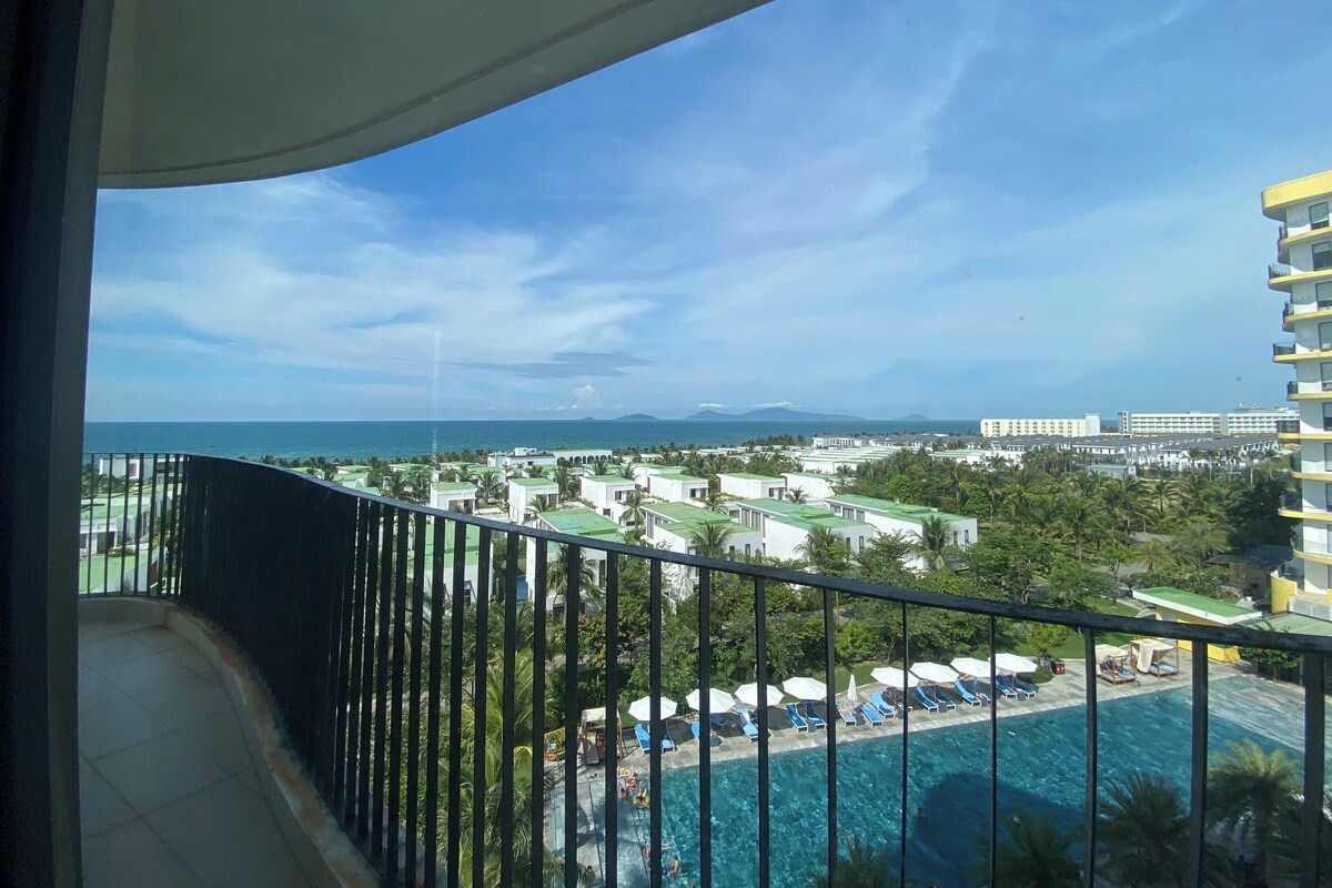 A balcony with a view of a swimming pool, palm trees, and distant buildings.