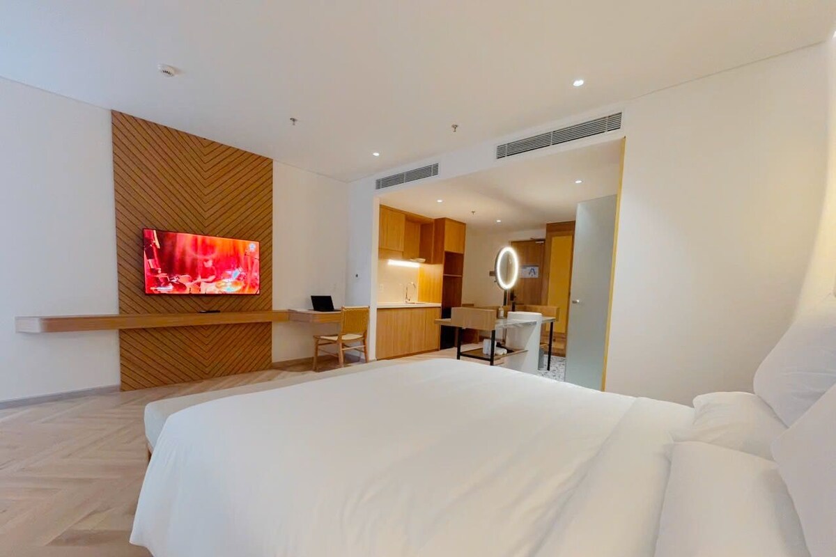 Een moderne hotelkamer met een groot bed, een televisie bevestigd aan een houten wandpaneel, een bureau met stoel, en een badkamer zichtbaar door een geopende deur.