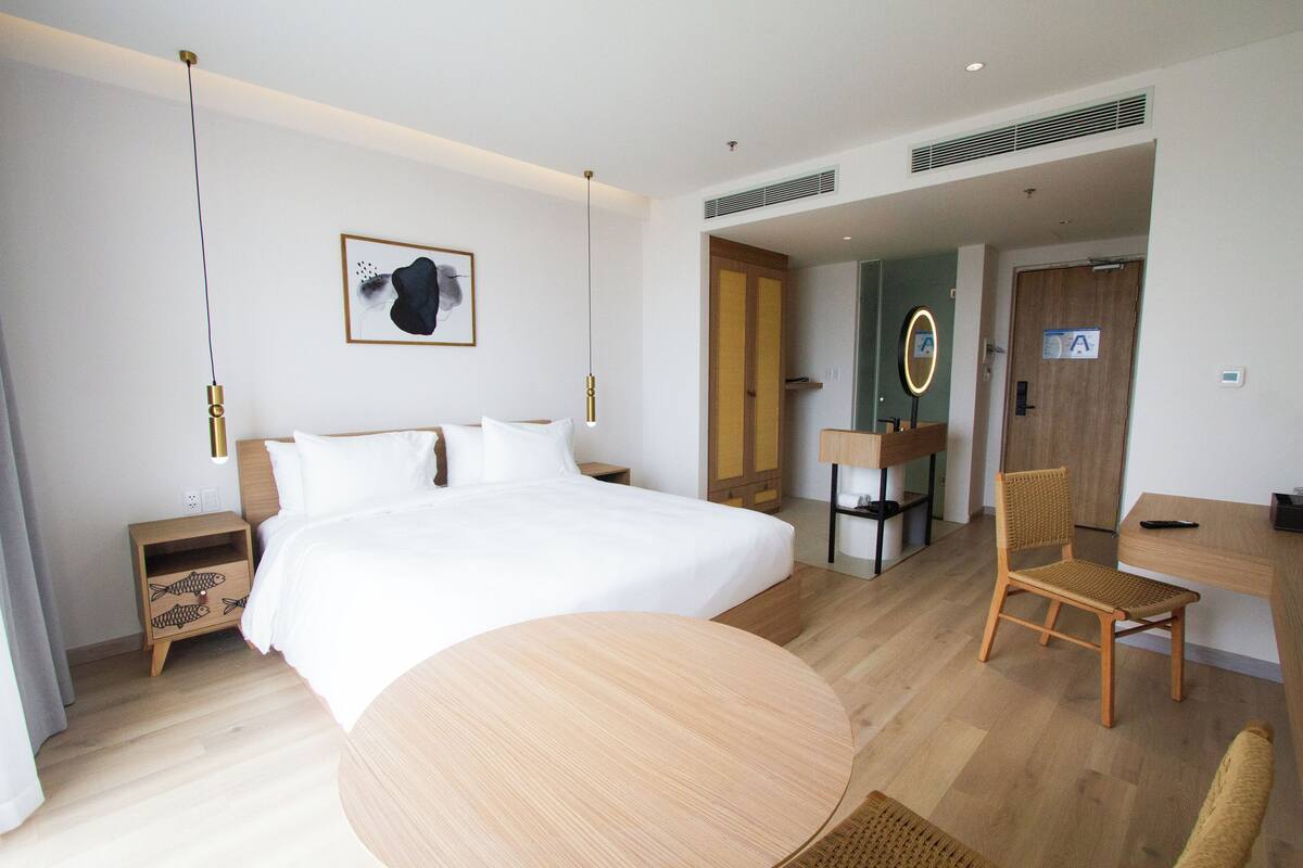 Habitación de hotel moderna con una cama grande, una mesa redonda de madera, un escritorio y una silla. Hay una mesita de noche con un diseño de pez, un espejo montado en la pared y una puerta que conduce a otra habitación.