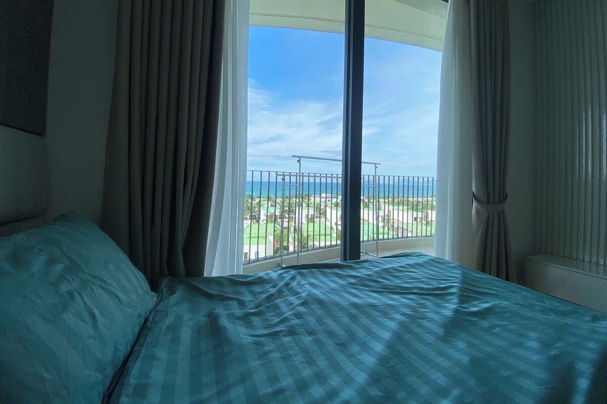 A hotel room with a bed, a window with curtains, and a view of the sea and greenery.
