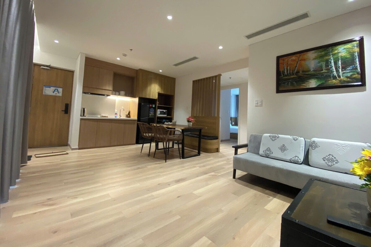A modern living room with a sofa, a dining area with chairs, a kitchen with wooden cabinets, and a painting on the wall.