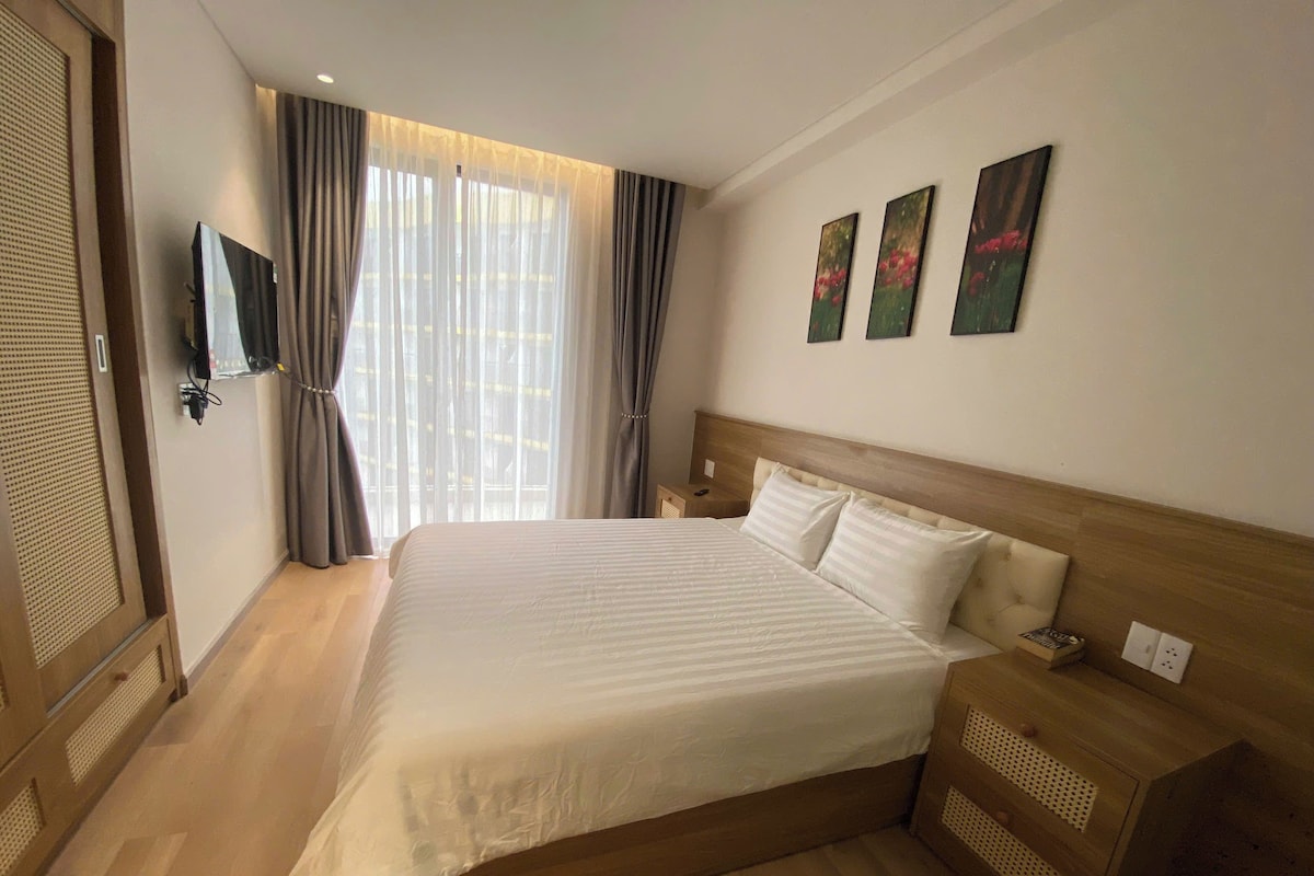 A hotel room with a large bed, a wooden headboard, a nightstand, a TV mounted on the wall, and a window with curtains.