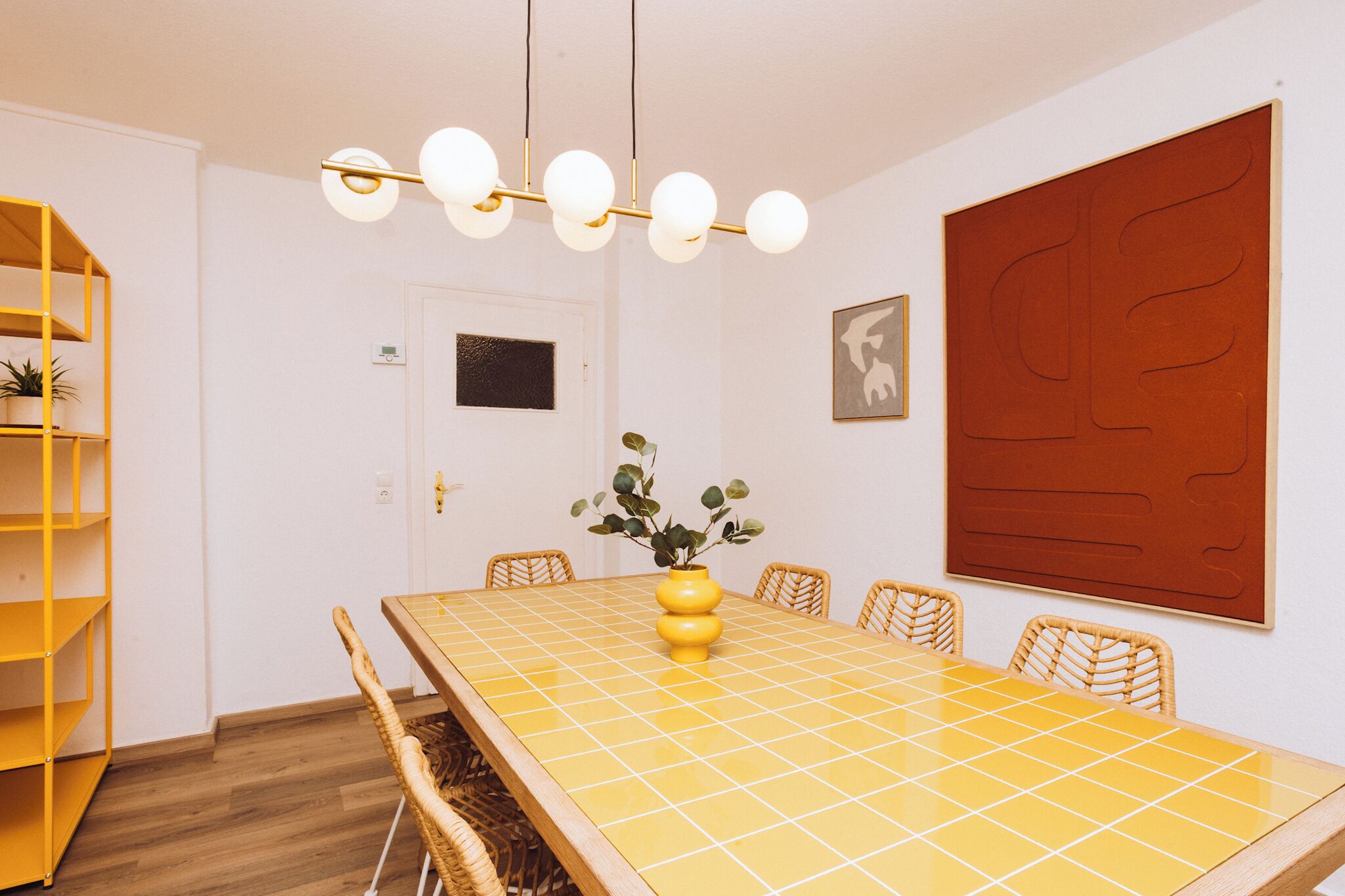 Une salle de conférence avec une table recouverte de carreaux jaunes, des chaises en osier, un vase jaune contenant des plantes, un luminaire suspendu moderne et une grande œuvre d’art abstraite accrochée au mur.