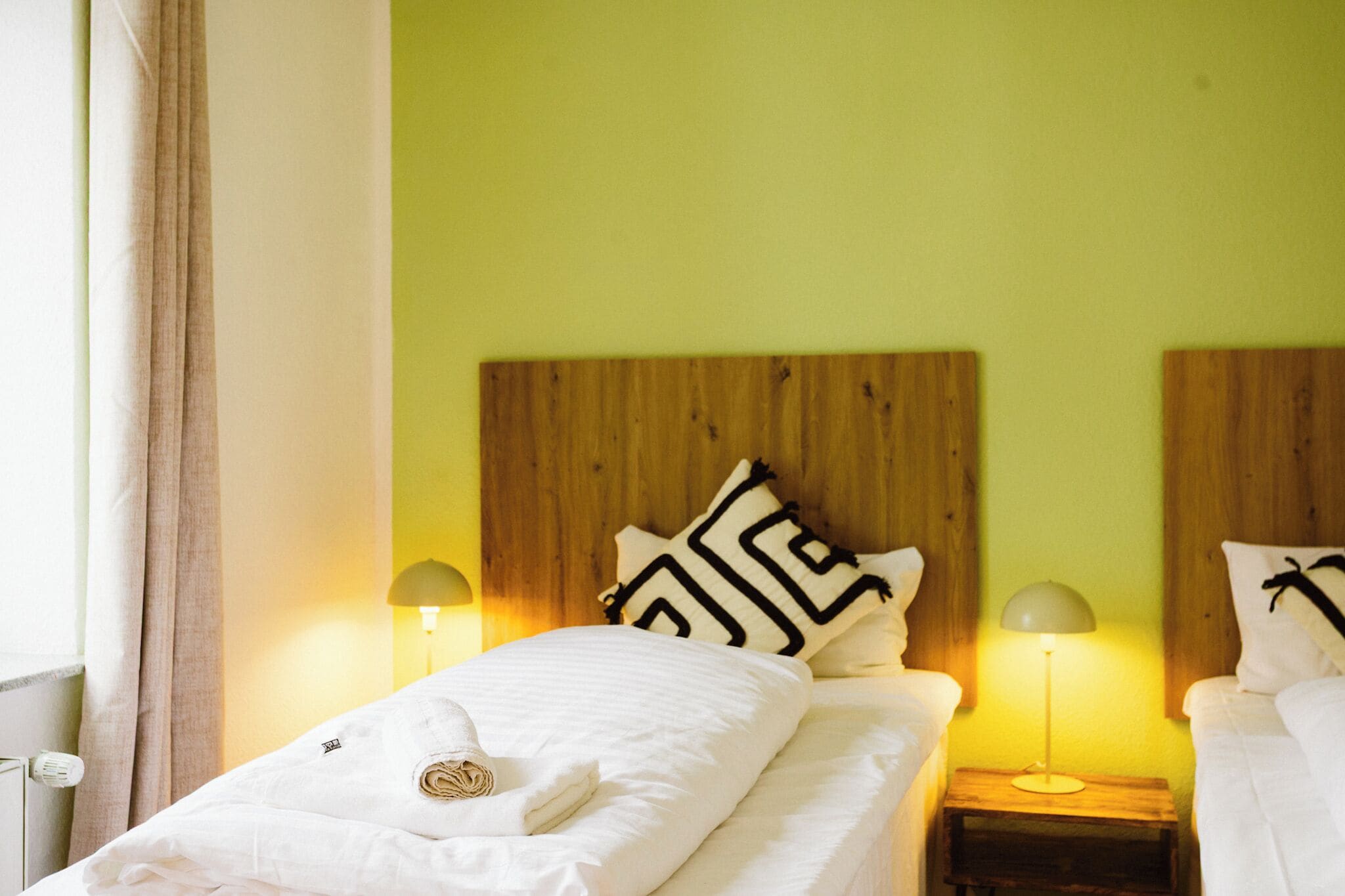 A hotel room with a wooden headboard, two bedside lamps, a bed with white bedding, and a window with curtains.