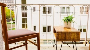 Ein Balkon mit Holzstuhl und kleinem Tisch, einer Topfpflanze und einem weißen Gebäude im Hintergrund.