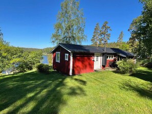 Une petite cabane rouge au toit en pente, située dans une zone herbeuse, avec un lac et des arbres en arrière-plan.