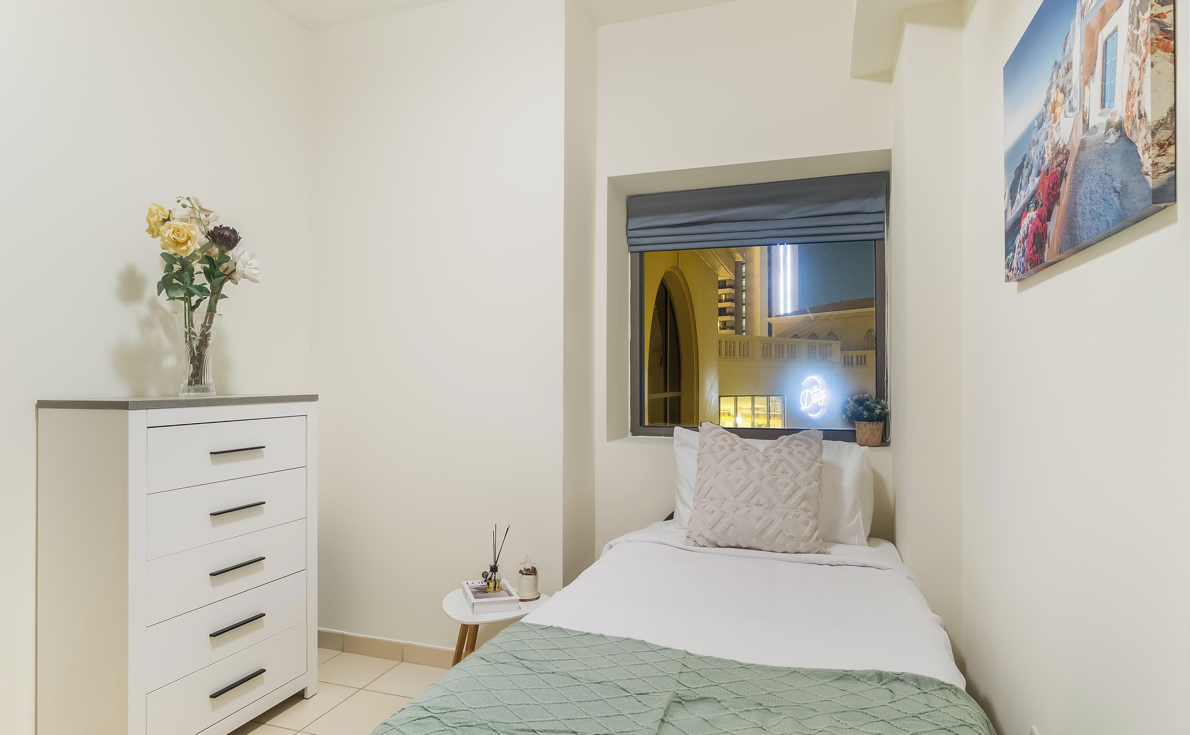 Une chambre à coucher avec un lit, une commode, une fenêtre donnant sur des immeubles et un tableau accroché au mur.