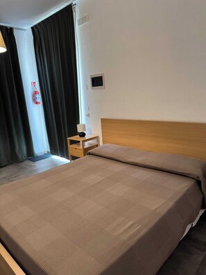 Quarto de hotel com cama, mesa de cabeceira, abajur, extintor de incêndio e porta com cortinas.