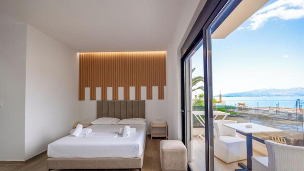 Ein modernes Hotelzimmer mit einem großen Bett, einem Holz-Kopfteil, einem Balkon mit Blick auf das Meer und die Berge sowie einer Glastür zum Balkon.