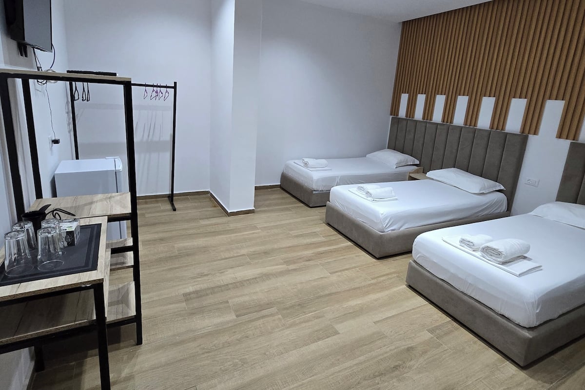 Habitación de hotel con tres camas, un escritorio con vasos, un televisor y una pared de paneles de madera.