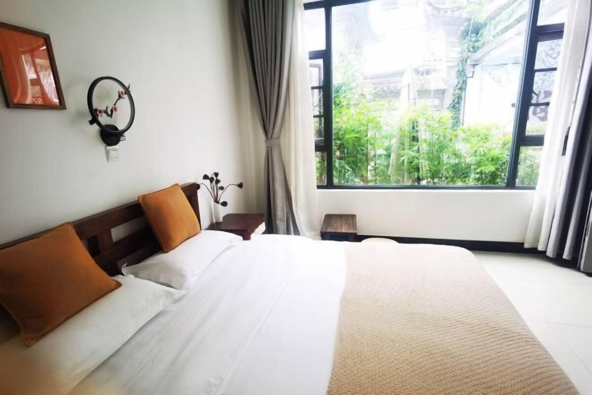 A hotel room with a large bed, a nightstand, a lamp, a wall-mounted clock, and a window with a view of greenery outside.