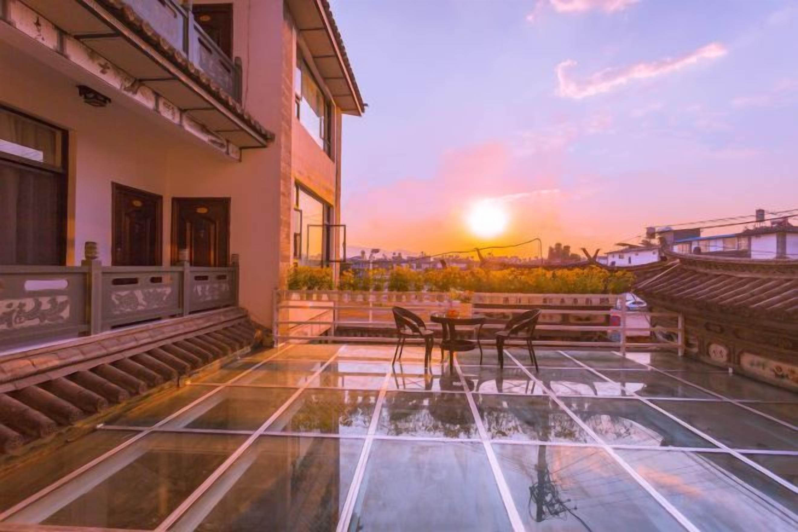 A rooftop terrace with a glass floor, a table, and chairs, overlooking a cityscape at sunset.
