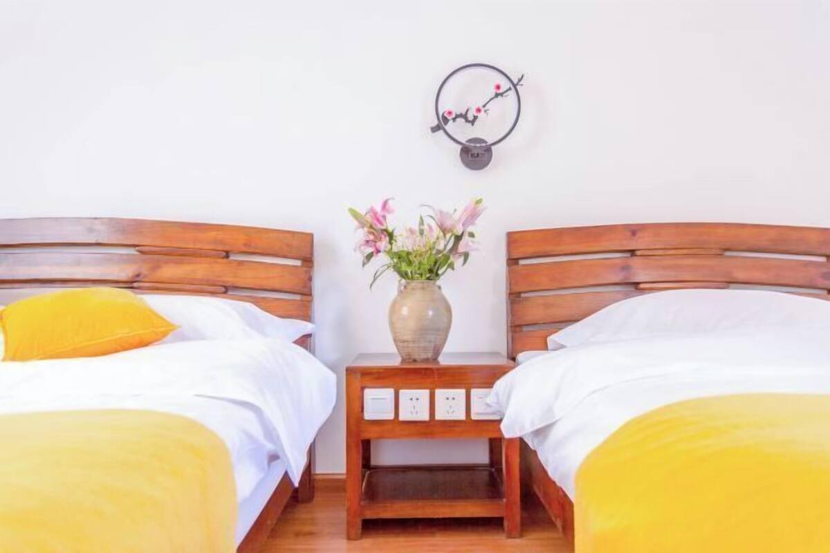 A double room with two wooden beds, each with a yellow bedspread and matching pillows. There is a small wooden nightstand between the beds with a vase of flowers and a wall clock.