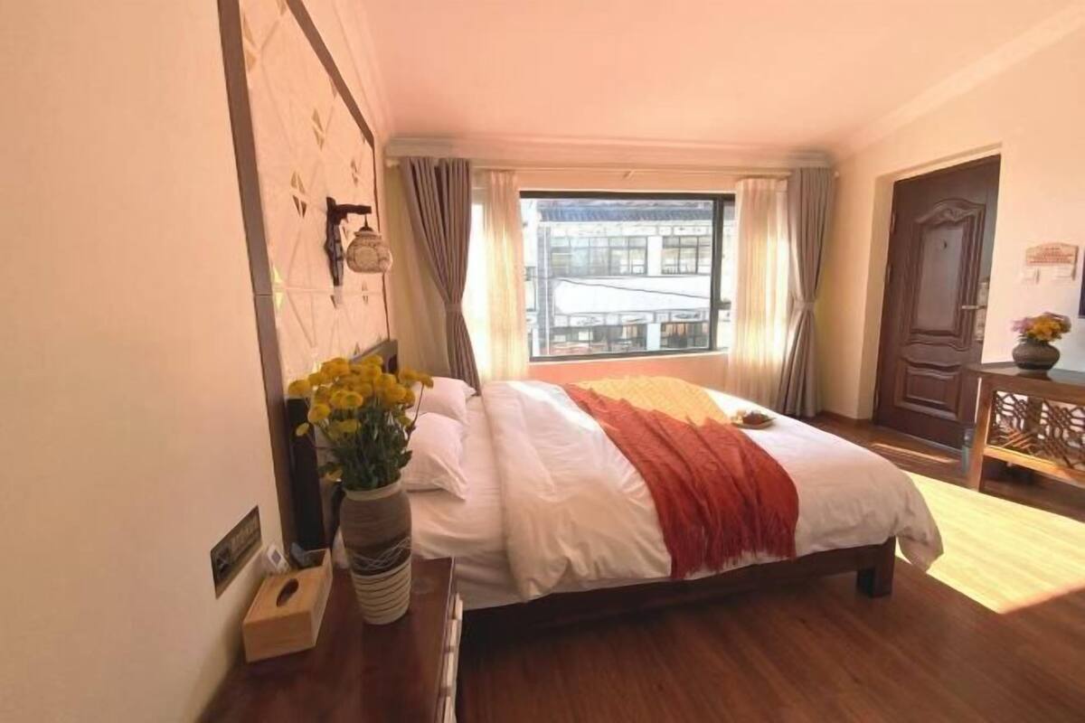 A hotel room with a bed, a window with curtains, a wooden desk, and a vase with flowers.
