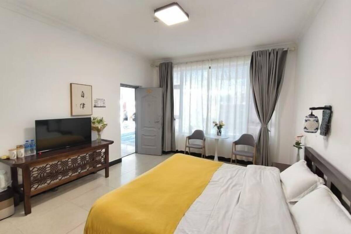 A hotel room with a bed, a TV on a wooden console, a small table with chairs, and a window with curtains.