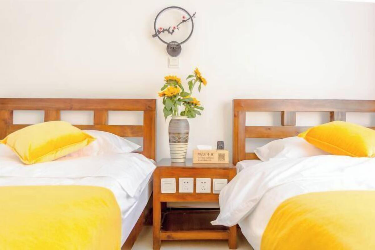 A hotel room with two beds, each with a wooden headboard, white bedding, and yellow pillows. There is a nightstand with a vase of sunflowers and a small sign.