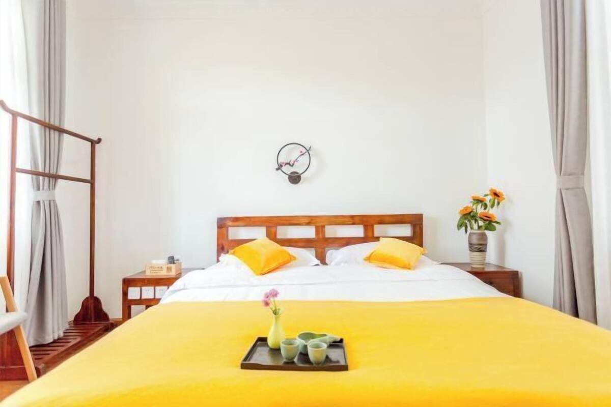 A hotel room with a bed, a bedside table, a nightstand, a wall clock, and a window with curtains.