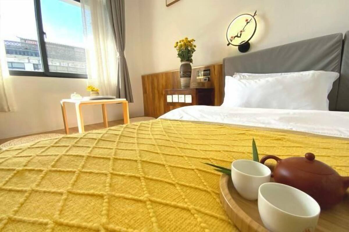 A hotel room with a bed, a bedside table with a vase of flowers, a clock, and a window with curtains.