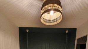 Un lit bien fait, recouvert d’une couverture vert foncé, deux tables de chevet avec des lampes et un grand luminaire suspendu au plafond, fait de fibres tressées.