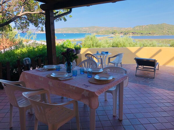 An outdoor dining area with a table set for two, chairs, a bench, and a view of the sea and hills.