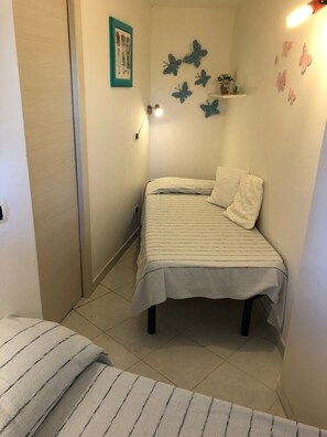 A small, neatly arranged bedroom with a bed, a bedside table, a chair, and a wall-mounted shelf with decorative items.