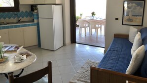 A compact kitchen with a refrigerator, a dining table with chairs, a bed with a blue-striped headboard, and a window with a view of the outdoors.