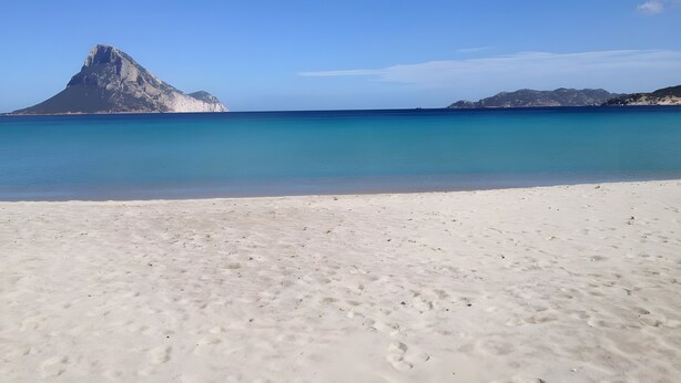 A beach with clear water, a mountain, and a sandy shore.