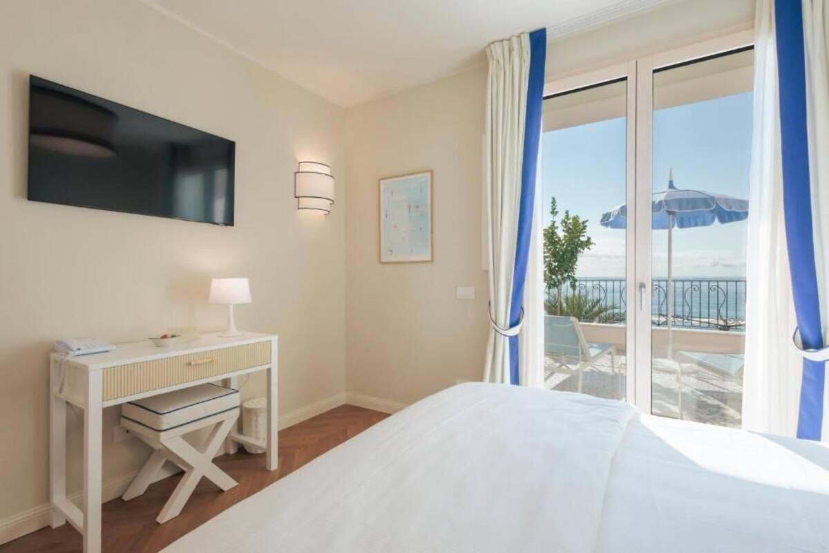 Una habitación de hotel con cama, un escritorio con lámpara, un cuadro y un balcón con vistas al mar.