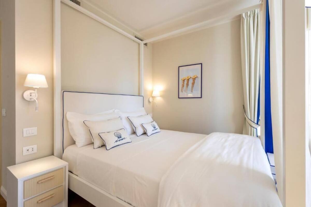 Una cama bien tendida con sábanas blancas y almohadas decorativas, una mesita de noche con lámpara y un cuadro en la pared.