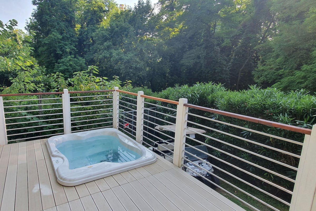 A hot tub on a wooden deck with a white railing, surrounded by lush greenery.