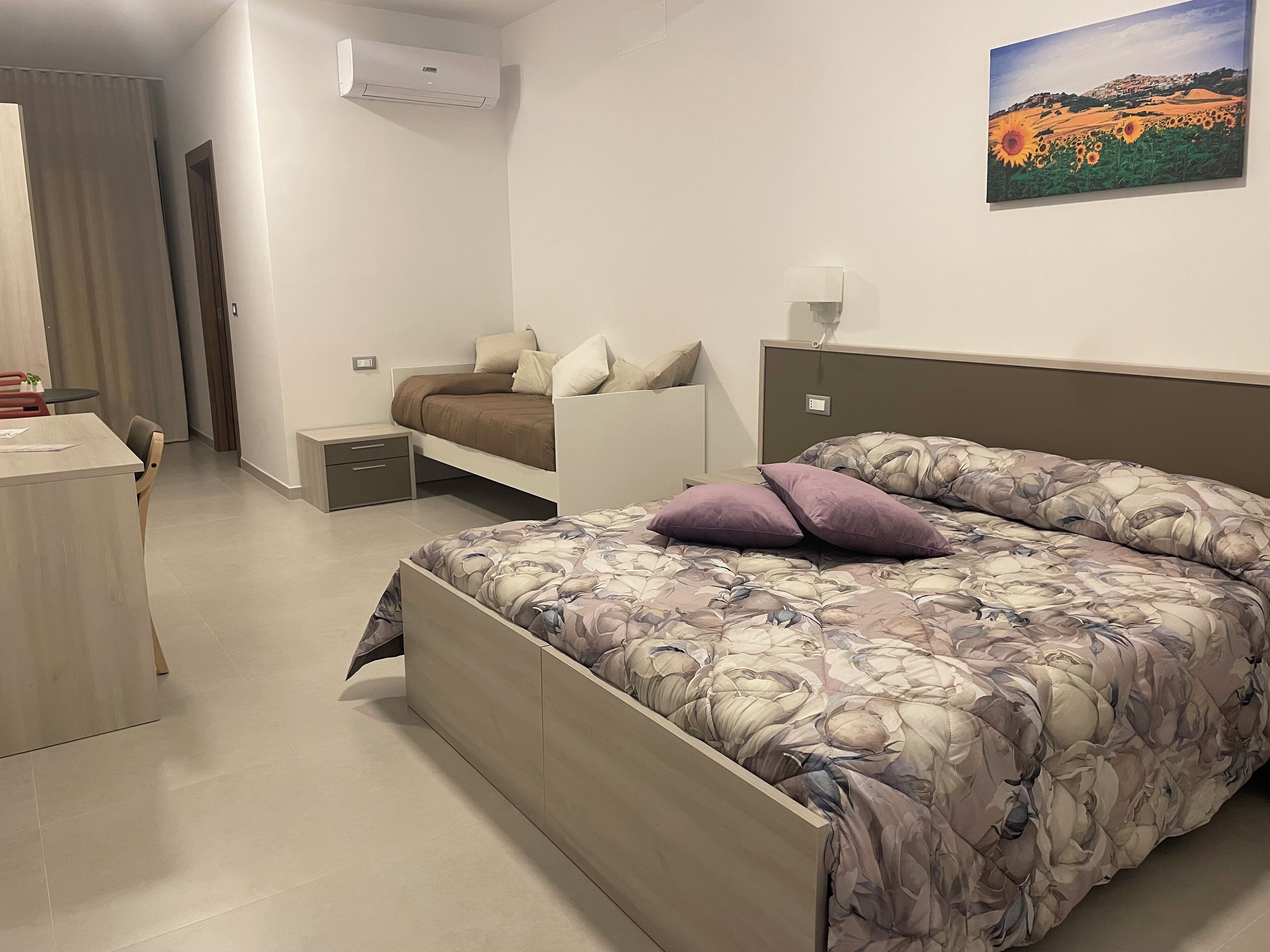 A bedroom with a bed, a desk, a chair, a nightstand, and a wall-mounted air conditioner.